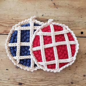 May include: Two round, crocheted coasters designed to resemble fruit pies. One coaster features a deep blue center with a cream-colored lattice crust, while the other has a bright red center with the same cream-colored lattice crust design.