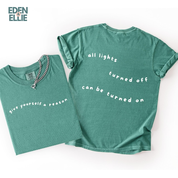 May include: Two teal-colored t-shirts with white text. One shirt reads "give yourself a reason", and the other says "all lights turned off can be turned on". A silver chain necklace is on one shirt. The shirts are from Eden & Ellie.