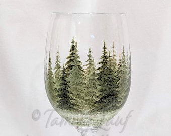May include: A clear glass wine glass featuring a hand-painted forest scene with various shades of green pine trees encircling the bowl. The base of the glass is clear and circular. The text tammypalette.com is visible on the glass surface.