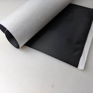 May include: A roll of white paper with a black sheet partially unrolled. The black sheet has a shiny surface and is partially folded over. The paper is on a white surface.