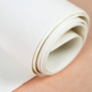 Puede incluir: Primer plano de un rollo de papel blanquecino. El papel está enrollado firmemente, revelando los bordes de las hojas. El papel tiene una textura suave y un tono ligeramente cálido.