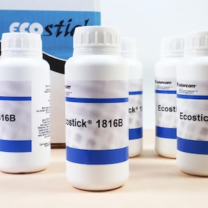 May include: Several white plastic bottles with blue stripes and white caps are arranged on a light-colored surface. The bottles have labels with the text "Costick 1816B" and other text. A box with the text "Ecostick" is in the background.