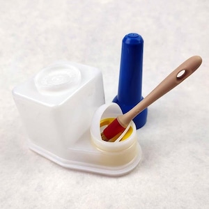 May include: A white plastic container with a clear lid, a blue plastic object, and a small container with a red and tan brush. The brush is angled towards the small container. The objects are on a white surface.