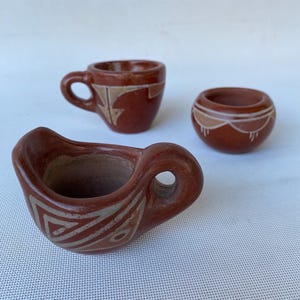 May include: Three small, handmade terracotta pottery pieces. The pieces include a small cup with a handle, a small bowl, and a small cup. Each piece has a reddish-brown base color with cream-colored geometric designs.
