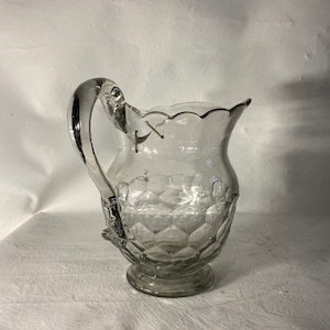 May include: A clear glass pitcher with a diamond pattern and a scalloped rim. The pitcher has a handle and a round base.