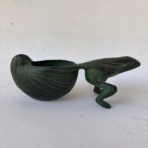 May include: A bronze-colored sculpture of a frog holding a nautilus shell. The shell forms a bowl, and the frog's body extends to support it. The sculpture has a weathered, antique appearance, with green and brown tones.