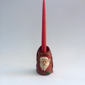May include: A red candle sits in a ceramic candle holder shaped like Santa Claus's face. The holder is brown with a white beard and red hat.