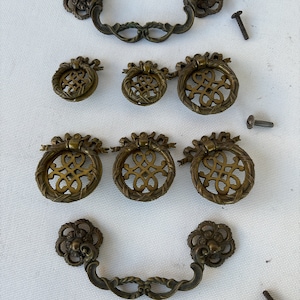 May include: A set of ten antique brass cabinet knobs and pulls with intricate floral and scroll designs. The knobs are round with a decorative center, and the pulls have a curved shape with a decorative end.