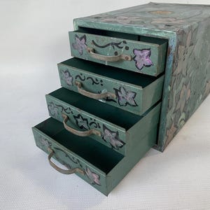 May include: A vintage-style, four-drawer storage box in shades of green, adorned with floral and leaf patterns. Each drawer features a metal handle and is decorated with purple flower accents. The box is a charming piece of decor.