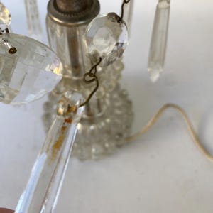 May include: A vintage clear glass lamp with hanging crystal accents. The lamp features a decorative base and a floral-shaped top. The crystals are various shapes and sizes, and the lamp has a cord for power.