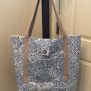May include: A blue and white paisley patterned tote bag with tan straps and a small pocket on the front. The bag has a button closure.