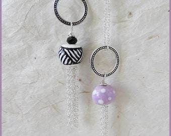 May include: Two silver necklaces with black and white patterned beads and a purple bead with white polka dots. Each necklace has a silver circle pendant and a silver tassel.