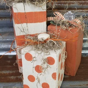 Puede incluir: Tres bloques decorativos de madera rústica. Uno está pintado de blanco con rayas naranjas, otro es naranja y el tercero es blanco con lunares naranjas. Todos están adornados con musgo seco y cuerda naranja.