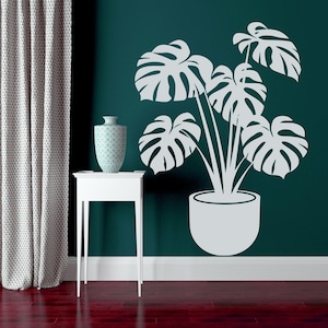 May include: A white silhouette of a potted monstera plant on a teal wall. The plant is in a white pot and has large, leafy leaves. A white side table with a vase on top is in the foreground.