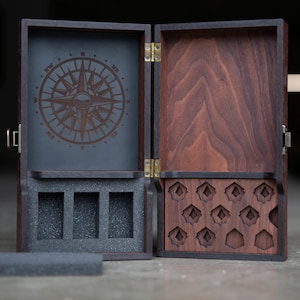 May include: A wooden box with a compass design on the lid and a compartment for dice. The box is open and the interior is lined with black foam. The box has a brass hinge and clasp.