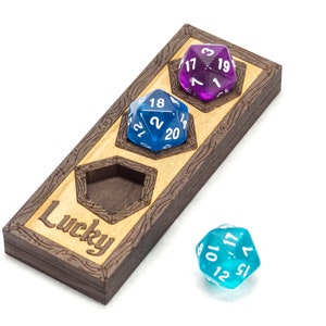 May include: A wooden dice tray with three slots for dice. The tray is engraved with the word "Lucky". There are three dice in the slots, one blue, one purple, and one turquoise.