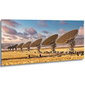May include: A panoramic view of a series of large white radio telescope satellite dishes arranged in a line across a dry, golden-brown desert landscape under a sky filled with soft, pink and blue clouds at sunset or sunrise.