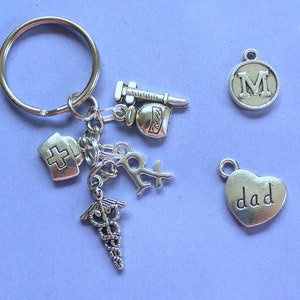 May include: Silver keychain with multiple charms, including a medical symbol, a pill bottle, a syringe, a cross, and a heart with the word "dad" on it. There is also a charm with the letter "M" on it.