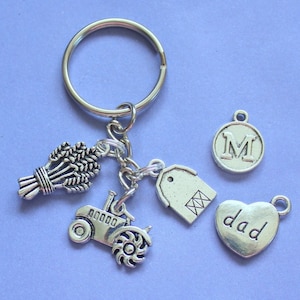 May include: Silver-coloured keyring with farming-themed charms. Features a tractor, wheat bundle, tag, initial "M" charm, and a heart charm with the word "dad". The charms are attached to a large metal ring.