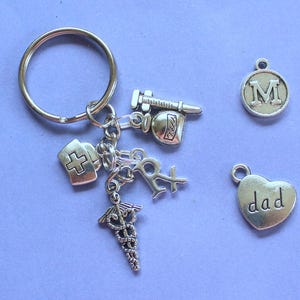 May include: Silver keychain with multiple charms, including a cross, a heart with the word "dad" on it, a bottle of pills, a syringe, and a letter "M".