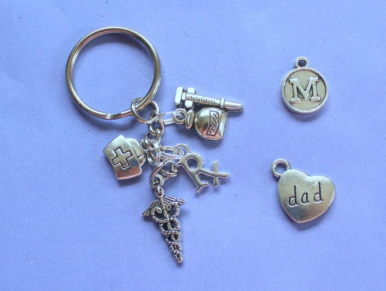 May include: Silver keychain with multiple charms, including a cross, a heart with the word "dad" on it, a bottle of pills, a syringe, and a letter "M".