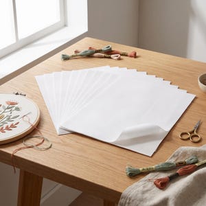 May include: A stack of white rectangular transfer paper sheets positioned centrally, accompanied by an embroidery hoop with a floral design, small gold scissors, and several skeins of green and peach embroidery floss arranged nearby.