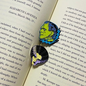 May include: Two enamel pins featuring classic monster designs. One pin depicts Frankenstein's monster in green and blue, the other shows the Bride of Frankenstein in black, yellow, and purple. Both pins are set against an open book.