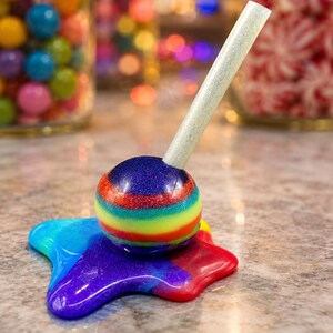 May include: A spherical rainbow-colored lollipop with a glittery finish, appearing to be melted into a star-shaped puddle of vibrant colors on a surface. A single, straight, light-colored stick extends upward from the center of the round candy.
