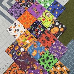 Puede incluir: Una colección de 25 cuadrados de tela con varios patrones de temática de Halloween. Los cuadrados están dispuestos en un patrón diagonal, con cada cuadrado presentando un diseño diferente, incluyendo calabazas, murciélagos, fantasmas y arañas. La tela es una mezcla de colores, incluyendo naranja, negro, morado, verde y blanco.
