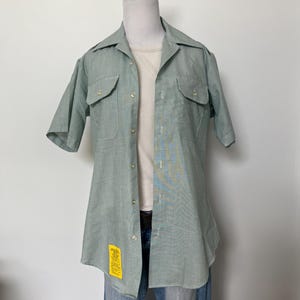 May include: A short-sleeved, light green button-down shirt with a pointed collar, two chest pockets with buttoned flaps, and a small yellow rectangular tag with black text attached to the lower front hem, worn over a plain white undershirt.