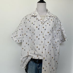 May include: A short-sleeved white button-down shirt featuring a repeating pattern of small, circular motifs in shades of brown and grey. The shirt has a classic collar, a single chest pocket, and is worn tucked into a pair of blue denim jeans.