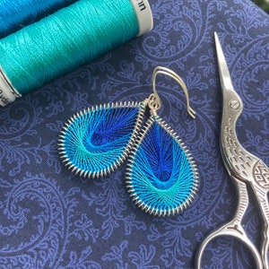 May include: A pair of blue and turquoise thread wrapped earrings with silver hoops. The earrings are on a dark blue fabric background with a pair of silver scissors.