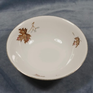 May include: White ceramic bowl with a gold rim and a delicate brown and pink leaf design. The bowl has a simple, elegant shape, perfect for serving soups or salads. A classic piece for any table setting.