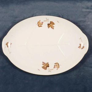 May include: An oval, off-white serving platter with a gold rim and leaf design. The platter features a central vein design and is decorated with brown and gold autumn leaves. The handles are on either side.