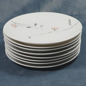 May include: A stack of eight white ceramic plates. Each plate features a delicate design of a branch with pink flowers and gray accents. The plates are round and have a simple, elegant aesthetic.