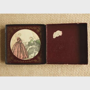 May include: An open, antique box with a maroon interior and a decorative, circular image. The image depicts two women in period dresses, one in pink and the other in green. The box has a dark exterior with gold filigree.