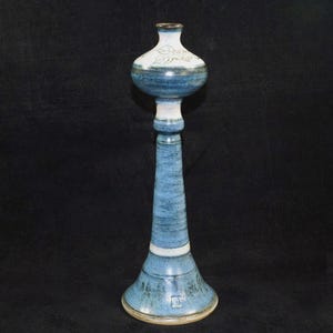 May include: A tall, handcrafted ceramic candlestick in shades of blue and white. The candlestick features a bulbous top with leaf designs, a slender, textured stem, and a flared base. The overall height is approximately 12 inches.
