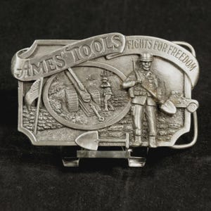 May include: Silver-toned metal belt buckle with the text "AMES TOOLS" and "FIGHTS FOR FREEDOM". The buckle features a soldier holding a shovel, a cannon, and an American flag. The buckle is rectangular with a curved top.