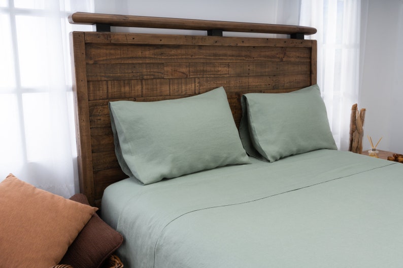 May include: A sage green linen bed sheet set with two matching pillowcases. The bed has a rustic wooden headboard with a metal accent.