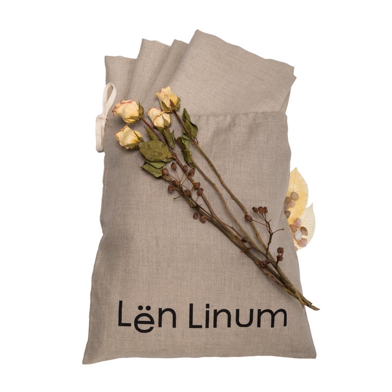 May include: A beige linen bag with a drawstring closure. The bag is filled with folded linen fabric and has the text "L&euml;n Linum" printed on the front. Dried flowers and leaves are placed on top of the bag.