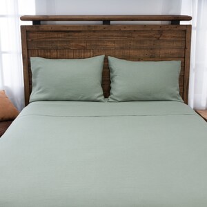 May include: A light green linen bed sheet with two matching pillowcases on a wooden bed frame. The bed is made with a brown and orange throw pillow.