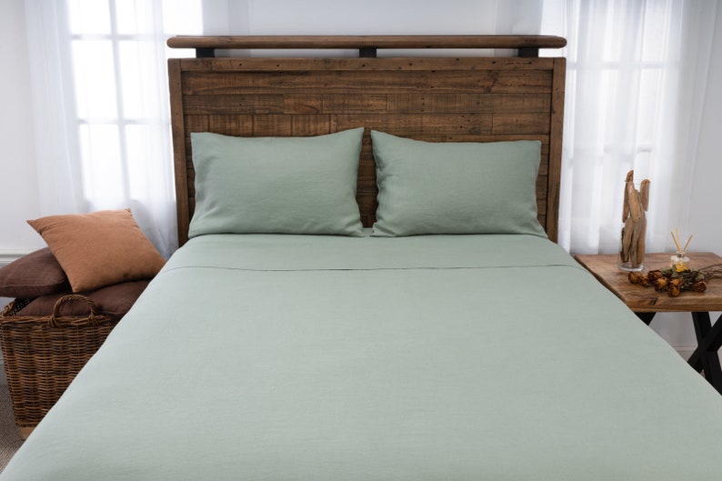 May include: A light green linen bed sheet with two matching pillowcases on a wooden bed frame. The bed is made with a brown and orange throw pillow.