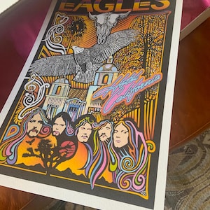 May include: A rectangular poster featuring the word EAGLES at the top, a central eagle and skull illustration, and the text Hotel California. The artwork includes five stylized portraits, a desert landscape with a tree, and architectural elements.