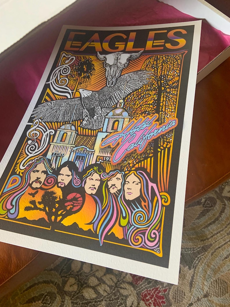 May include: A rectangular poster featuring the word EAGLES at the top, a central eagle and skull illustration, and the text Hotel California. The artwork includes five stylized portraits, a desert landscape with a tree, and architectural elements.