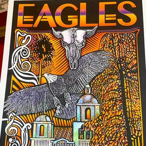 May include: A rectangular concert poster featuring the word EAGLES in a gradient orange and purple font. The artwork displays a central eagle in flight, a steer skull, a building labeled Troubadour 1976, and swirling white musical notes.