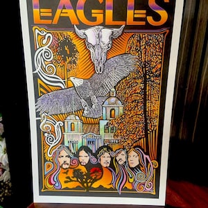May include: A rectangular poster featuring the word EAGLES in orange and purple gradient lettering. The artwork displays a white animal skull, a flying eagle, a building labeled Troubadour 1976, and five stylized portraits with flowing hair.