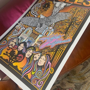 May include: A rectangular concert poster featuring the band Eagles and the text Hotel California. The artwork displays a bald eagle, a skull, and the band members with colorful, swirling hair against a black background with orange and yellow accents.