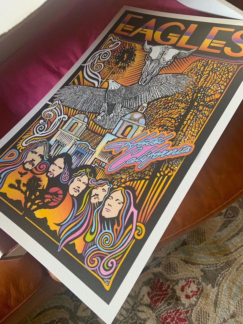 May include: A rectangular concert poster featuring the band Eagles and the text Hotel California. The artwork displays a bald eagle, a skull, and the band members with colorful, swirling hair against a black background with orange and yellow accents.