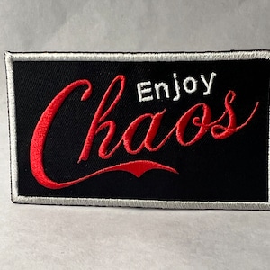 Può includere: Una toppa ricamata nera con un bordo bianco. La toppa presenta un testo corsivo rosso che dice "Enjoy Chaos".