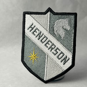 Può includere: Una toppa ricamata grigia e bianca con un bordo nero. La toppa ha la forma di uno scudo e presenta la parola "Henderson" in lettere bianche e una testa di lupo stilizzata in grigio. Una stella dorata si trova nell'angolo inferiore sinistro.